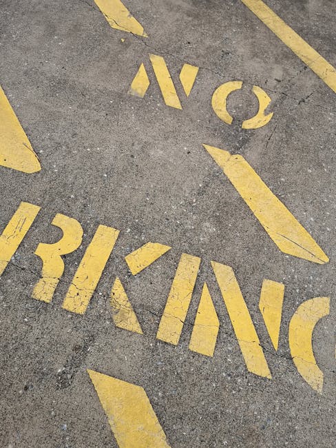 Close-up view of yellow parking bay suspension markings painted on an asphalt surface outside a property, with the words 'NO PARKING' visible alongside directional arrows. The markings are slightly worn, with some areas showing minor fading and chipping. The surrounding pavement appears rough with small cracks and textured stones. In the background, part of a loading area near the property entrance can be seen, where a man with a van from Man With a Van Morden might be engaged in home relocation activities, such as loading furniture, boxes, and packaging materials. The outdoor setting reflects the preparation stage of a furniture transport or moving process, emphasizing the importance of parking restrictions for smooth removal logistics. Such markings help facilitate efficient loading and unloading during house removals and are relevant for ensuring compliance with local parking regulations for Morden removals services.
