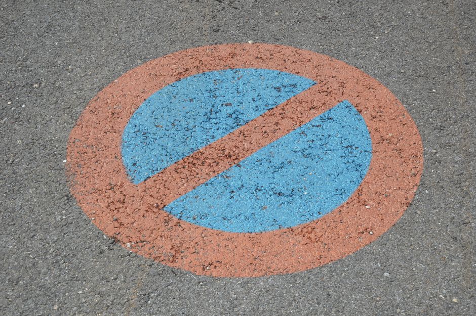 A circular no parking zone marking painted on an asphalt surface, featuring a blue inner circle with a diagonal red line through it, indicating a parking restriction. The marking is situated on a paved area outside a residential property, which may be used for loading or unloading during a house removal. The surrounding pavement appears worn and textured, with the marking clearly visible and adhering to standard road safety signage. The image context suggests it could be relevant for relocation services or removals logistics, such as ensuring compliance with parking restrictions during a home relocation undertaken by Man With a Van Morden, which operates in the Morden area.