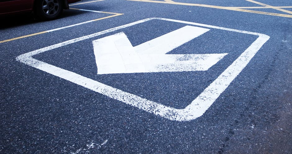 Close-up view of yellow parking bay suspension markings painted on an asphalt surface outside a property, with the words 'NO PARKING' visible alongside directional arrows. The markings are slightly worn, with some areas showing minor fading and chipping. The surrounding pavement appears rough with small cracks and textured stones. In the background, part of a loading area near the property entrance can be seen, where a man with a van from Man With a Van Morden might be engaged in home relocation activities, such as loading furniture, boxes, and packaging materials. The outdoor setting reflects the preparation stage of a furniture transport or moving process, emphasizing the importance of parking restrictions for smooth removal logistics. Such markings help facilitate efficient loading and unloading during house removals and are relevant for ensuring compliance with local parking regulations for Morden removals services.