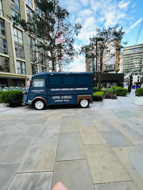 A small, vintage-style blue van with a sliding side door is parked on a paved urban area outside modern apartment buildings. The van features white text advertising London Dock, fresh coffee, and packaging boxes, suggesting its use for delivery or promotional purposes. The vehicle is in the process of being loaded or unloaded, with part of a black loading ramp extended from the back of the van onto the pavement. Tall residential buildings with numerous windows, balconies, and modern architecture surround the scene, along with green bushes and trees lining the walkway. The area appears well-maintained, with natural daylight illuminating the scene, and the van's compact size indicating it is suitable for furniture transport or small home relocation tasks, as part of a house removals service offered by Man With a Van Morden.