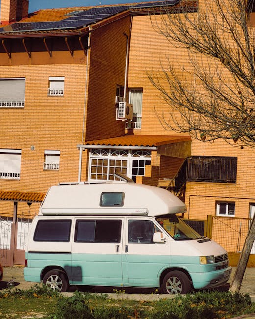 A light blue and white campervan parked on the street in front of a multi-storey brick residential building during daylight. The vehicle is positioned parallel to the pavement, with its side doors closed and windows visible. The building features several small windows with white shutters, an external air conditioning unit attached to a balcony, and a black staircase leading to an upper entrance. A tree with bare branches is situated beside the van, partially obscuring the building façade. The scene reflects a typical home relocation setting, with the van likely used for furniture transport and packing during house moving or removals, as part of the services offered by Man With a Van Morden.