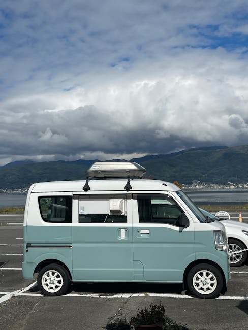 A small, vintage-style blue van with a sliding side door is parked on a paved urban area outside modern apartment buildings. The van features white text advertising London Dock, fresh coffee, and packaging boxes, suggesting its use for delivery or promotional purposes. The vehicle is in the process of being loaded or unloaded, with part of a black loading ramp extended from the back of the van onto the pavement. Tall residential buildings with numerous windows, balconies, and modern architecture surround the scene, along with green bushes and trees lining the walkway. The area appears well-maintained, with natural daylight illuminating the scene, and the van's compact size indicating it is suitable for furniture transport or small home relocation tasks, as part of a house removals service offered by Man With a Van Morden.