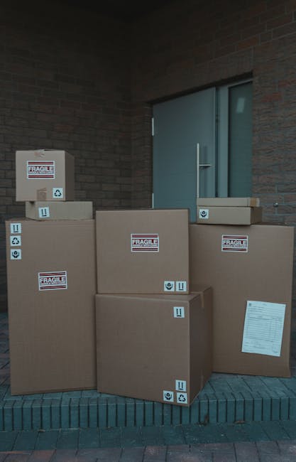 A collection of large cardboard boxes stacked outside a building entrance on a brick paved area, with some boxes labeled 'FRAGILE' and marked with handling symbols such as arrows, a fragile icon, and a recycling symbol. The boxes vary in size and are arranged in front of a modern brick wall and a grey metal door with a vertical handle. The environment appears to be part of a home relocation process, supported by visible packing materials and labels indicating careful handling of household items during furniture transport or moving services. The outdoor lighting is neutral, and the scene captures the loading process of packed belongings prepared for removal or delivery, consistent with the services offered by Man With a Van Morden.