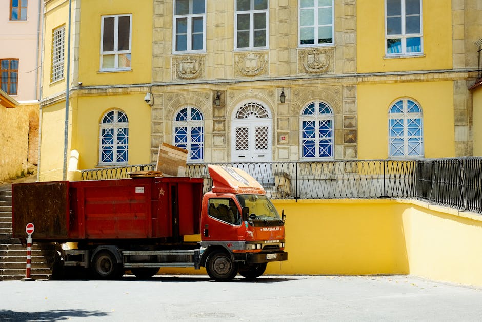 A red moving truck with a closed cubic cargo area is parked on a paved street in front of a yellow, multi-story building with ornate stone window frames and arched windows fitted with white lattice-patterned security bars. The truck is positioned close to a yellow retaining wall with a black metal railing on top, adjacent to outdoor stairs leading up to a side entrance of the building. Cardboard boxes and flat packing materials are placed on top of the truck and appear to be ready for a home relocation process. The scene is well-lit with natural daylight, and the truck is aligned parallel to the building, suggesting the beginning of the furniture transport and packing process typical of professional removals services such as those offered by Man With a Van Morden.