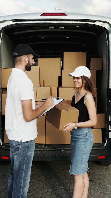 A man with a beard wearing a black cap, white t-shirt, and jeans is inside a van, taking notes on a clipboard. He is next to a woman wearing a white cap, black sleeveless top, and denim skirt, who is holding a medium-sized cardboard box. They are surrounded by several other packed cardboard boxes, some stacked inside the van. The van has an open rear door revealing a spacious interior filled with moving boxes placed on the floor and against the walls, ready for home relocation or furniture transport. The scene is outdoors on a paved surface, with natural lighting illuminating the area, and the surroundings include a background with greenery and a clear sky. This image demonstrates packing and loading processes in professional removals, representing the services provided by Man With a Van Morden.
