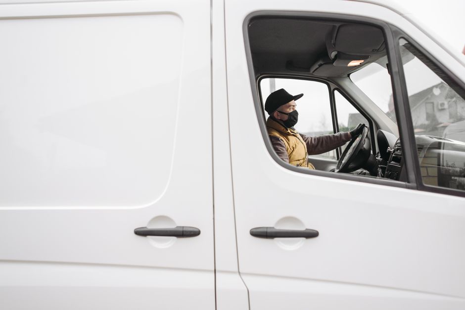 Close-up side view of a white commercial van parked outdoors, with the driver's side door open, revealing a man wearing a black face mask, black cap, and brown vest sitting at the wheel. The driver is inside the vehicle, holding the steering wheel, and appears to be preparing for a home relocation or furniture transport. The interior of the van is visible through the window, and the background shows a cloudy sky and faint outlines of other vehicles or buildings. The scene is set in a typical loading area or driveway, consistent with the moving and removals services offered by Man With a Van Morden, including packing and loading processes for house removals.