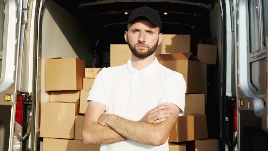 A man with a beard and wearing a black cap and white polo shirt stands inside a moving van with open side doors, arms crossed, in front of a large collection of cardboard boxes stacked on the vehicle'