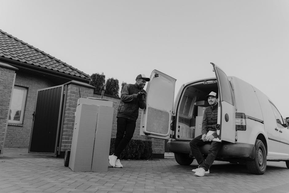 A man wearing a white t-shirt, grey pants, and a black cap is outside in daylight, loading three cardboard boxes into the side of a white commercial van parked on a suburban street. The man is holding