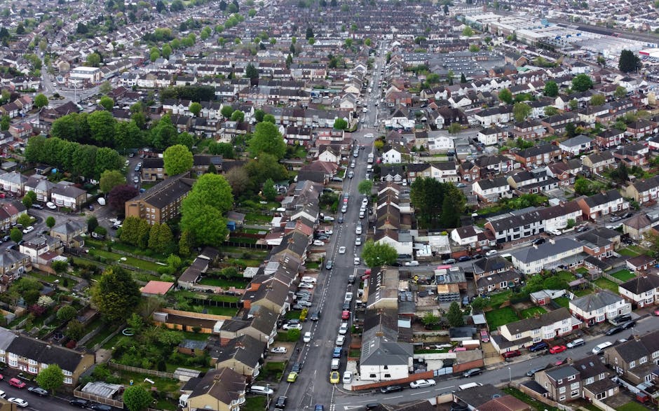 Aerial view of a residential neighbourhood with tightly packed houses and streets, featuring a mix of terraced and detached homes, along with several trees of varying sizes and shades of green. The streets are lined with parked cars, and some houses have small gardens or backyards. A narrow central road runs vertically through the image, with vehicles parked along both sides, and a few cars visible in motion. The surrounding area includes more densely arranged houses extending to the horizon, with urban infrastructure visible in the distance. The lighting indicates daytime, with overcast skies casting diffuse light. This scene illustrates the typical environment where a home relocation or furniture transport might occur, with residents possibly preparing for a move involving packing and loading items for transport, which companies like Man With a Van Morden facilitate.