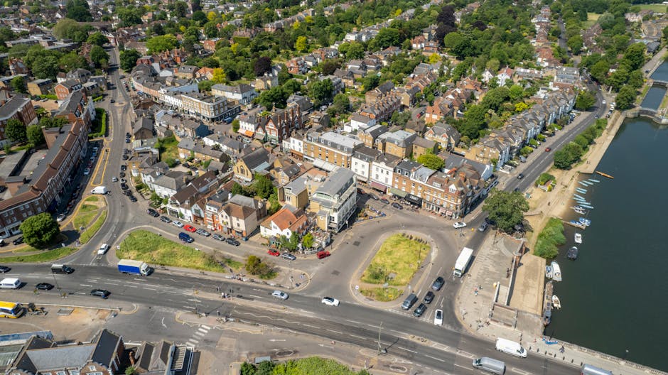 Aerial view of a residential area in Morden, showing a mix of terraced and semi-detached houses with varied roof styles and building materials, adjacent to a river with boats docked along a paved promenade. The scene includes a busy street with multiple lanes, cars, and delivery vans, some of which are parked on the roadside or in designated parking areas. Several green spaces and small parks are visible, along with a roundabout near the center of the image, surrounded by a few commercial buildings and shops. The environment appears well-lit with natural daylight, highlighting the urban layout and the close proximity of homes, roads, and waterfront amenities. This setting reflects a typical residential and commercial neighbourhood suitable for home relocation activities, with the presence of loading zones and accessible streets, relevant to services like house removals provided by Man With a Van Morden, especially during furniture transport and packing processes.