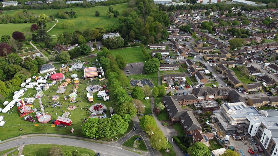 Aerial view of a residential neighbourhood with tightly packed houses and streets, featuring a mix of terraced and detached homes, along with several trees of varying sizes and shades of green. The streets are lined with parked cars, and some houses have small gardens or backyards. A narrow central road runs vertically through the image, with vehicles parked along both sides, and a few cars visible in motion. The surrounding area includes more densely arranged houses extending to the horizon, with urban infrastructure visible in the distance. The lighting indicates daytime, with overcast skies casting diffuse light. This scene illustrates the typical environment where a home relocation or furniture transport might occur, with residents possibly preparing for a move involving packing and loading items for transport, which companies like Man With a Van Morden facilitate.