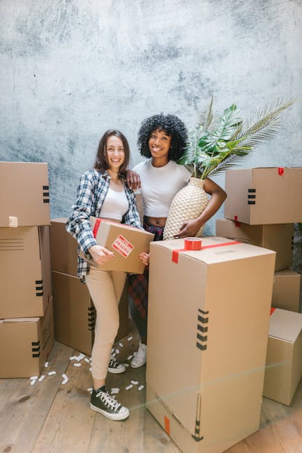 Two women are standing indoors among several unpacked cardboard moving boxes, some sealed with red tape, in what appears to be a home or storage area. One woman, with long brown hair and wearing a blue plaid shirt over a white top and beige pants, is holding a small cardboard box and smiling at the camera. The other woman, with curly black hair and wearing a white t-shirt and plaid skirt, is holding a large decorative vase with long green palm leaves. The boxes are stacked around them, some on the wooden floor, indicating they are in the process of packing or unpacking during a home relocation. The background features a textured wall with a neutral, light-coloured finish. The scene reflects furniture transport and packing activities typical of professional removals services, such as those by Man With a Van Morden, supporting efficient house move logistics.