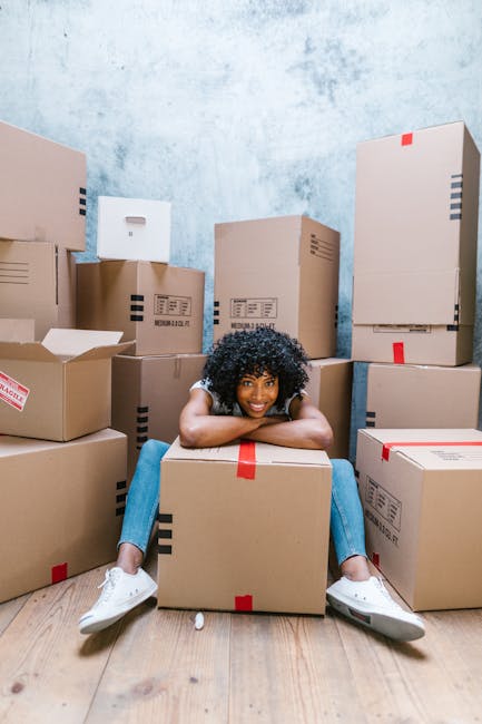 A young woman with curly black hair, dressed casually in a white top, blue jeans, and white sneakers, is sitting on the wooden floor inside a room filled with stacked cardboard moving boxes of various sizes. She is smiling and resting her arms on a large box in front of her. Several boxes are wrapped with red and black tape, some are partially open revealing packing materials such as bubble wrap and tissue paper. The room has a light blue textured wall in the background, indicating an indoor setting prepared for home relocation. The scene depicts the packing and moving process, with boxes arranged around her, and a focus on the logistics involved in house removals, as managed by a professional service like Man With a Van Morden.