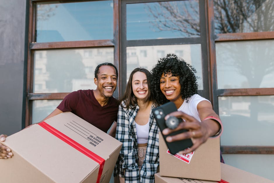 Two women are standing indoors among several unpacked cardboard moving boxes, some sealed with red tape, in what appears to be a home or storage area. One woman, with long brown hair and wearing a blue plaid shirt over a white top and beige pants, is holding a small cardboard box and smiling at the camera. The other woman, with curly black hair and wearing a white t-shirt and plaid skirt, is holding a large decorative vase with long green palm leaves. The boxes are stacked around them, some on the wooden floor, indicating they are in the process of packing or unpacking during a home relocation. The background features a textured wall with a neutral, light-coloured finish. The scene reflects furniture transport and packing activities typical of professional removals services, such as those by Man With a Van Morden, supporting efficient house move logistics.