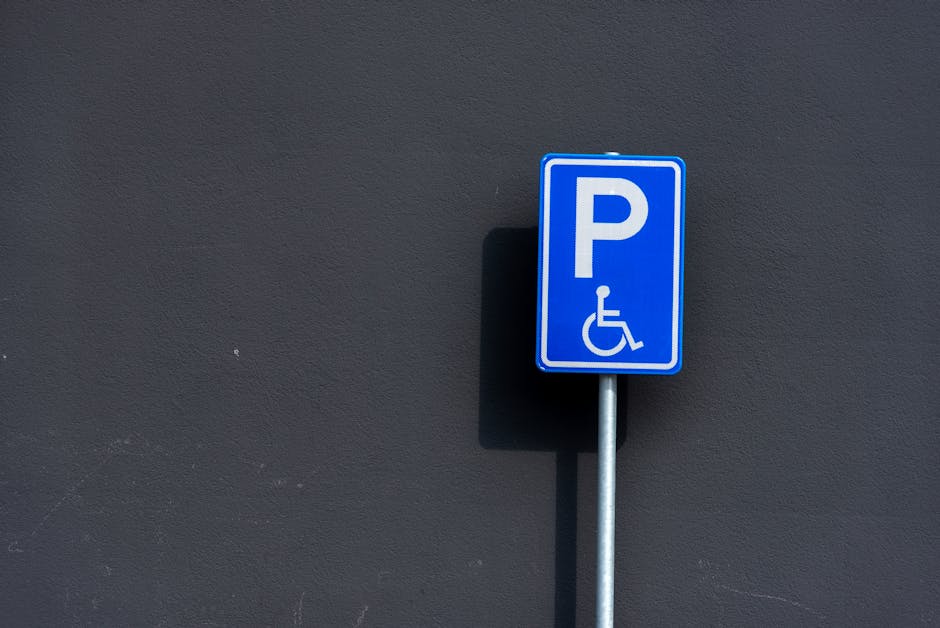 A blue rectangular parking sign with a white 'P' and a wheelchair symbol indicating accessible parking is mounted on a metal pole against a dark grey wall. The sign is positioned slightly to the right of center in the image, casting a small shadow onto the wall behind it. The environment appears to be an outdoor area adjacent to a property, suggesting a designated accessible parking space suitable for home relocation or furniture transport services. This signage setup supports parking and access considerations during moves, as provided by Man With a Van Morden, relevant to the context of house removals and moving logistics in Morden.