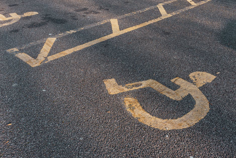 A blue rectangular parking sign with a white 'P' and a wheelchair symbol indicating accessible parking is mounted on a metal pole against a dark grey wall. The sign is positioned slightly to the right of center in the image, casting a small shadow onto the wall behind it. The environment appears to be an outdoor area adjacent to a property, suggesting a designated accessible parking space suitable for home relocation or furniture transport services. This signage setup supports parking and access considerations during moves, as provided by Man With a Van Morden, relevant to the context of house removals and moving logistics in Morden.