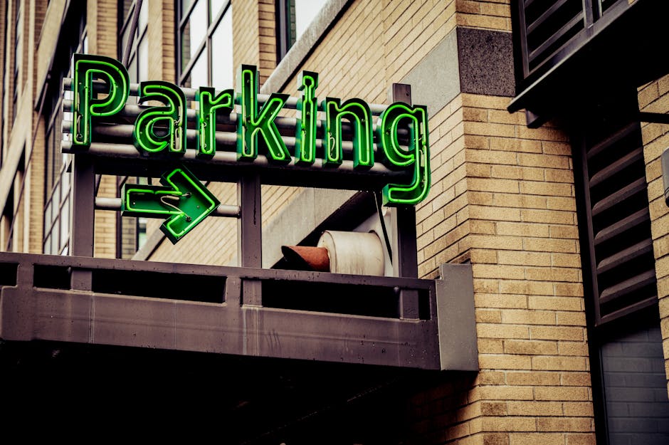 A neon green illuminated sign reading 'Parking' with an arrow pointing to the right is mounted on a metal frame attached to the exterior of a multi-storey building. The sign is positioned above a small balcony with a concrete railing, which holds a large beige flowerpot and some construction or packing materials such as cardboard or plastic wrapping. Behind the sign, the building's brickwork is visible, featuring a mixture of tan and brown bricks, and part of a window with black louvered shutters can be seen on the right side. The scene suggests an urban environment potentially related to house or office relocations, with the signage indicating available parking or loading zone, relevant for moving services by Man With a Van Morden.