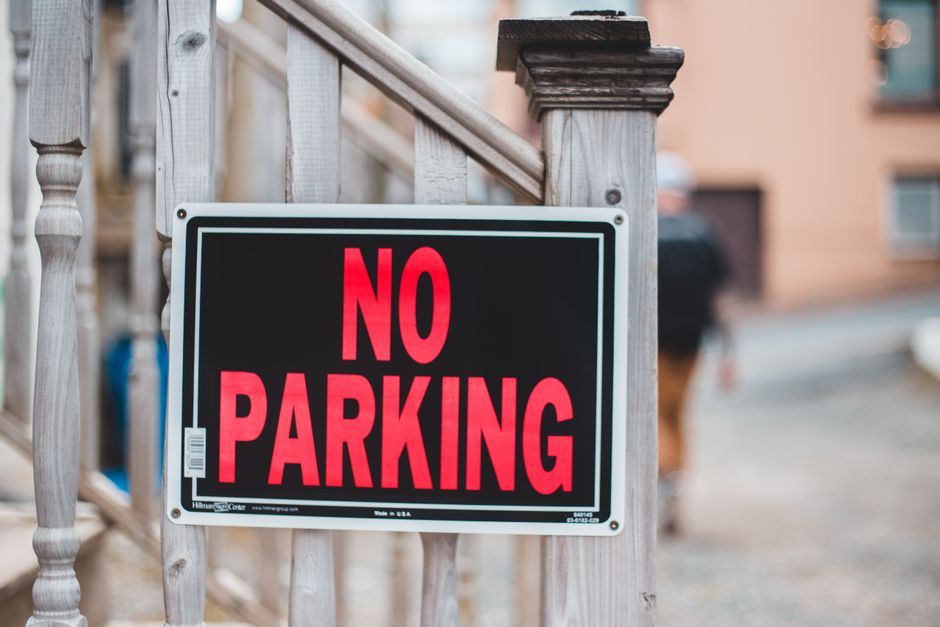 A black and white 'No Parking' sign with bold red lettering is mounted on a wooden railing outside a residential property in Morden. The sign is positioned on a staircase bannister, with the building's exterior visible in the background, including a blurred view of a driveway and parked cars. The surrounding environment appears to be a typical suburban street, with natural lighting and a clear focus on the sign. This setting reflects the typical parking restrictions encountered during home relocation or furniture transport, where parking suspensions or restrictions may affect the logistics of a house removal, as managed by Man With a Van Morden, a professional removals service.