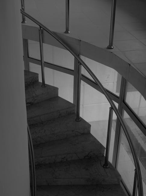 A curved marble staircase inside a building featuring a polished metal handrail supported by vertical metal balusters. The staircase is positioned near a glass partition with metal framing, allowing natural light to illuminate the area. Adjacent to the staircase, there is a section of tiled flooring, and part of a radiator is visible along the wall. The environment appears to be clean and well-maintained, with neutral tones and soft lighting, typical of a professional home or office relocation setting. This image relates to the process of moving household or office furniture, where careful handling and protective measures may be employed during a house removal or furniture transport, as suggested by the context of [PAGE_TITLE] and services provided by [COMPANY_NAME].