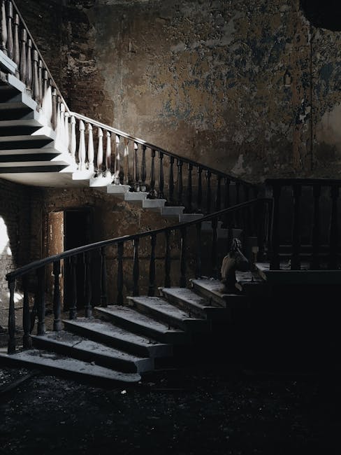 A worn, wooden staircase inside an old building features two sections connected by a landing, with white-painted steps and dark, aged railings on both sides. The upper section curves upward along the wall, while the lower section extends downward, with some steps showing signs of chipping and wear. The surrounding wall has peeling paint and exposed brick, indicating age and disrepair. Natural light enters from a nearby window, casting shadows across the stairs and highlighting the textured, distressed surfaces. This staircase is part of an interior intended for home or building renovation projects involving careful packing and moving, with Man With a Van Morden experienced in handling such interior stairway relocations to protect surfaces during furniture transport and home refurbishment processes.