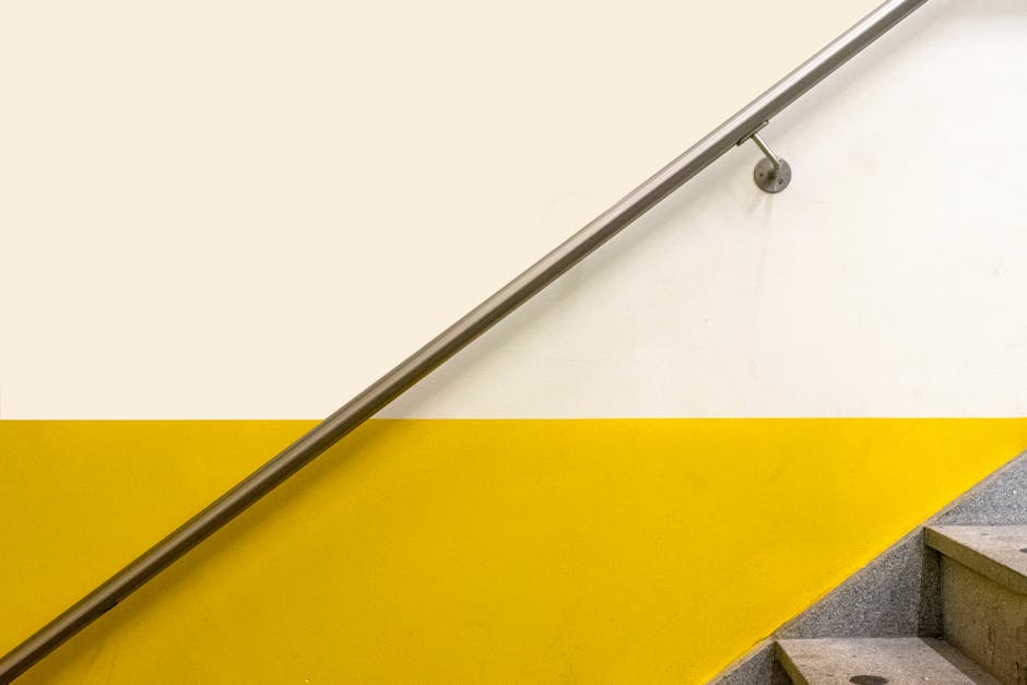 A curved marble staircase inside a building featuring a polished metal handrail supported by vertical metal balusters. The staircase is positioned near a glass partition with metal framing, allowing natural light to illuminate the area. Adjacent to the staircase, there is a section of tiled flooring, and part of a radiator is visible along the wall. The environment appears to be clean and well-maintained, with neutral tones and soft lighting, typical of a professional home or office relocation setting. This image relates to the process of moving household or office furniture, where careful handling and protective measures may be employed during a house removal or furniture transport, as suggested by the context of [PAGE_TITLE] and services provided by [COMPANY_NAME].