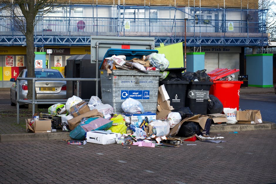 An overflowing collection of black, green, and red waste bins, along with a large grey mixed waste container filled with discarded cardboard, paper, plastic packaging, and household rubbish, is situated on a paved area outside a building. Some cardboard boxes and loose rubbish are scattered around the bins, spilling onto the pavement. In the background, there is a metal railing, a parked silver car with a visible license plate, and a retail storefront with signs indicating it is a fish bar and convenience store, with a blue scaffolding structure above the building. The scene is captured in daylight with natural lighting, illustrating typical waste disposal and rubbish collection processes associated with home relocation and moving logistics, which Man With a Van Morden handles as part of comprehensive removals services.
