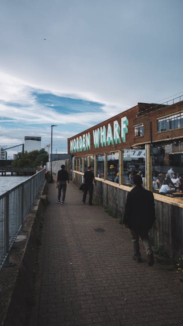 A group of people walking along a paved pathway beside Morden Wharf, a building with large glass windows and a prominent sign reading 'Morden Wharf' on its exterior. The pathway is bordered by a metal railing on the left and a wooden fence on the right, with some individuals dressed in dark clothing carrying no visible items. In the background, the sky is partly cloudy with patches of blue, and the scene suggests a busy, waterfront area. The image captures the environment around a home relocation or moving service site, with visible areas suitable for loading or unloading furniture or boxes as part of a house removals or packing and moving process, with the company Man With a Van Morden occasionally referenced.