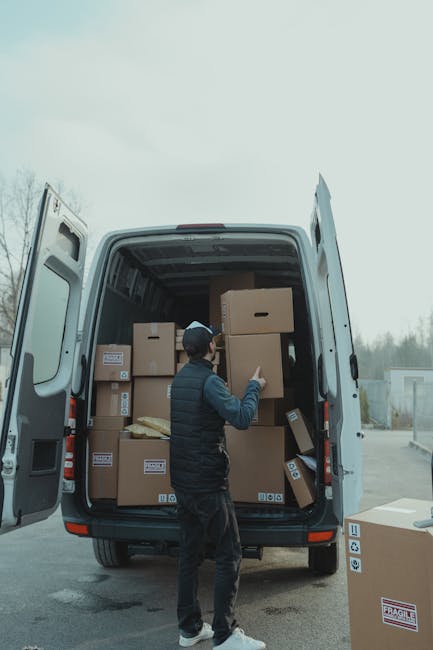 A man wearing a dark jacket, black trousers, and a cap is standing outside the open rear doors of a white moving van, which is parked on a paved driveway. Inside the van, numerous cardboard boxes of various sizes are neatly stacked, some secured with plastic wrap and others with labels visible on their sides. The boxes are positioned on a wooden floor base, with a larger box being lifted or moved by the man, indicating the process of furniture transport or packing during a home relocation. Several packing materials, including bubble wrap and packing paper, can be seen among the boxes. To the right of the van, on the pavement, there is an additional cardboard box, slightly tilted, that appears ready for loading or unloading. The background features a cloudy sky and a distant view of trees and a light industrial or storage area, consistent with an outdoor setting for moving services. This scene exemplifies the logistics involved in packing, moving, and loading household items as part of a professional removals service by Man With a Van Morden, emphasizing careful handling and organized transport for house removals or furniture transport processes.