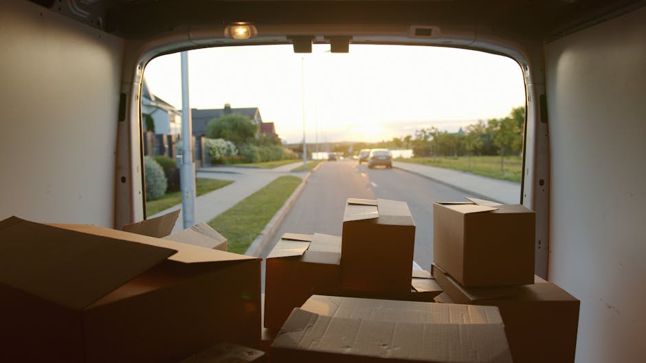View from inside a moving van showing several packed cardboard boxes, some with flaps open, arranged on the floor near the rear opening, ready for a house removal. The van is parked on the pavement of a residential street during sunset, with a clear sky, a few cars driving past, and houses with front gardens visible in the background. The interior of the van is plain, with neutral coloured walls, and the boxes are in various sizes, indicating a household packing and furniture transport process. This scene depicts part of a home relocation operation managed by Man With a Van Morden, highlighting the logistics involved in packing, loading, and transportation of belongings during a house move.
